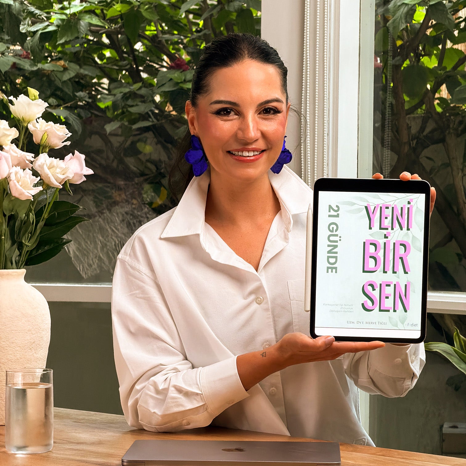 Woman holding a tablet with 'Yeni Bir Sen' on it, standing in a room with plants and a vase.