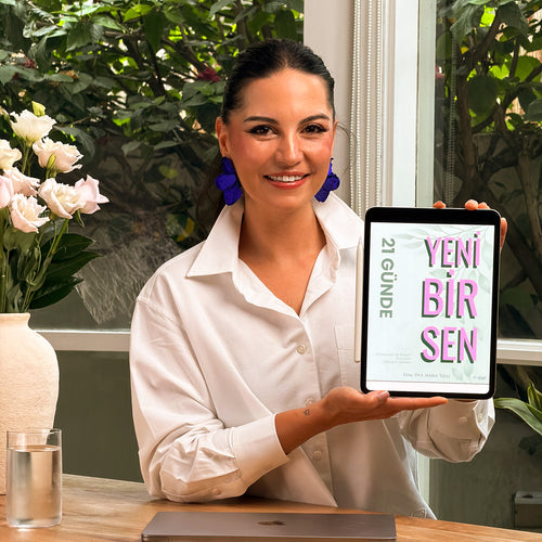 Woman holding a tablet with 'Yeni Bir Sen' on it, standing in a room with plants and a vase.